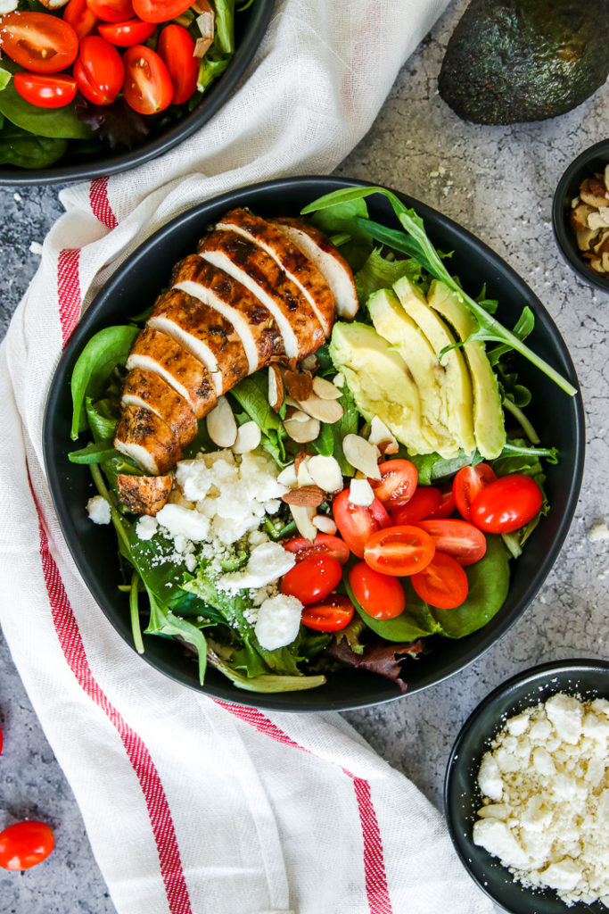 two black bowls of balsamic grilled chicken salad with feta, sliced almonds, tomatoes, and avocado