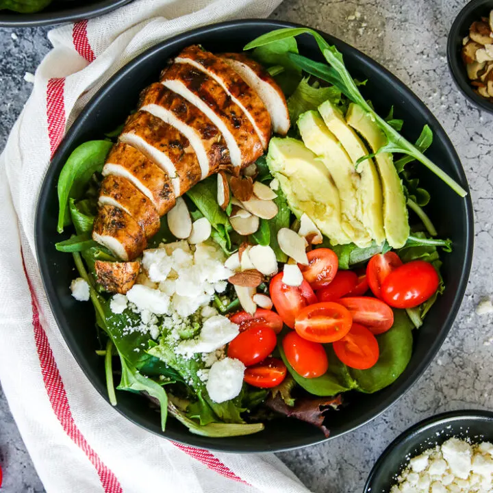 two black bowls of balsamic grilled chicken salad with feta, sliced almonds, tomatoes, and avocado