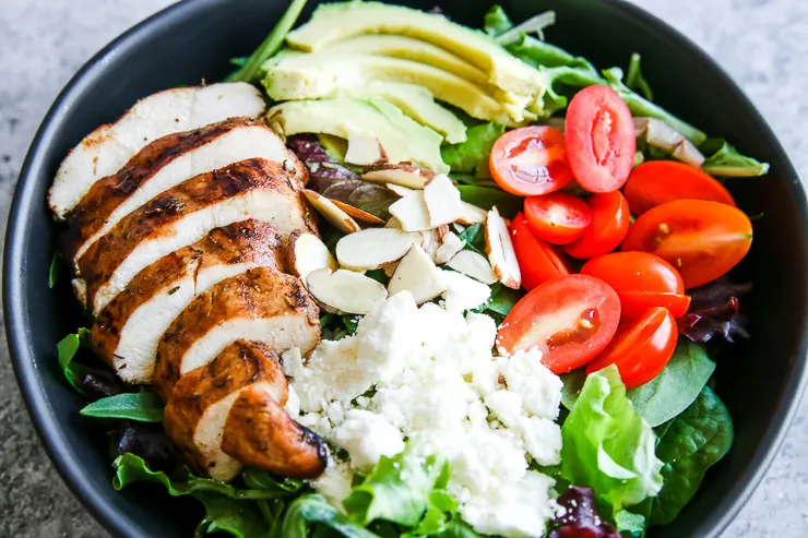 a close image of the sliced chicken breast, crumbled feta, slivered almonds, sliced cherry tomatoes, and sliced avocado on a spring mix salad