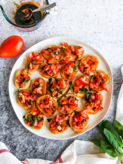 a plate of bruschetta with balsamic glaze next to a tomato, basil, and glass cup of balsamic glaze