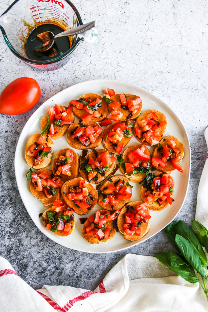 a plate of bruschetta with balsamic glaze next to a tomato, basil, and glass cup of balsamic glaze