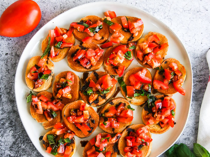 a plate of bruschetta with balsamic glaze next to a tomato, basil, and glass cup of balsamic glaze