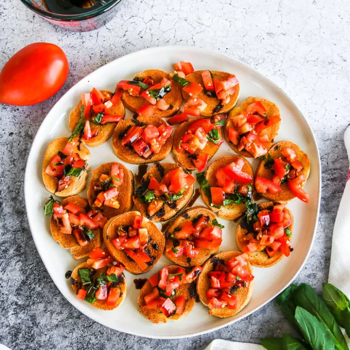 a plate of bruschetta with balsamic glaze next to a tomato, basil, and glass cup of balsamic glaze