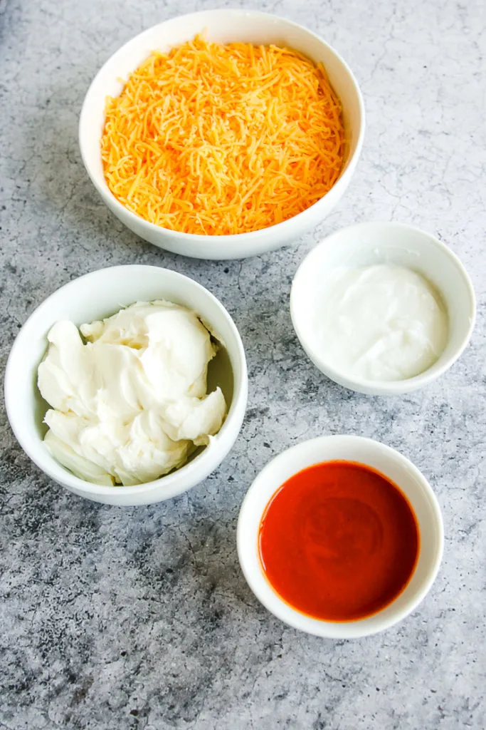 four bowls of shredded cheddar cheese, neufchatel cheese, greek yogurt, and buffalo sauce