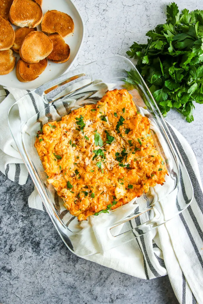 a glass baking dish full of baked buffalo cauliflower dip next to toasted bread and parsley