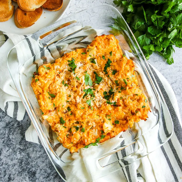 a glass baking dish full of baked buffalo cauliflower dip next to toasted bread and parsley