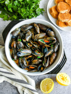 a saute pan full of garlic butter mussels next to parsley, toasted bread slices, and lemon halves