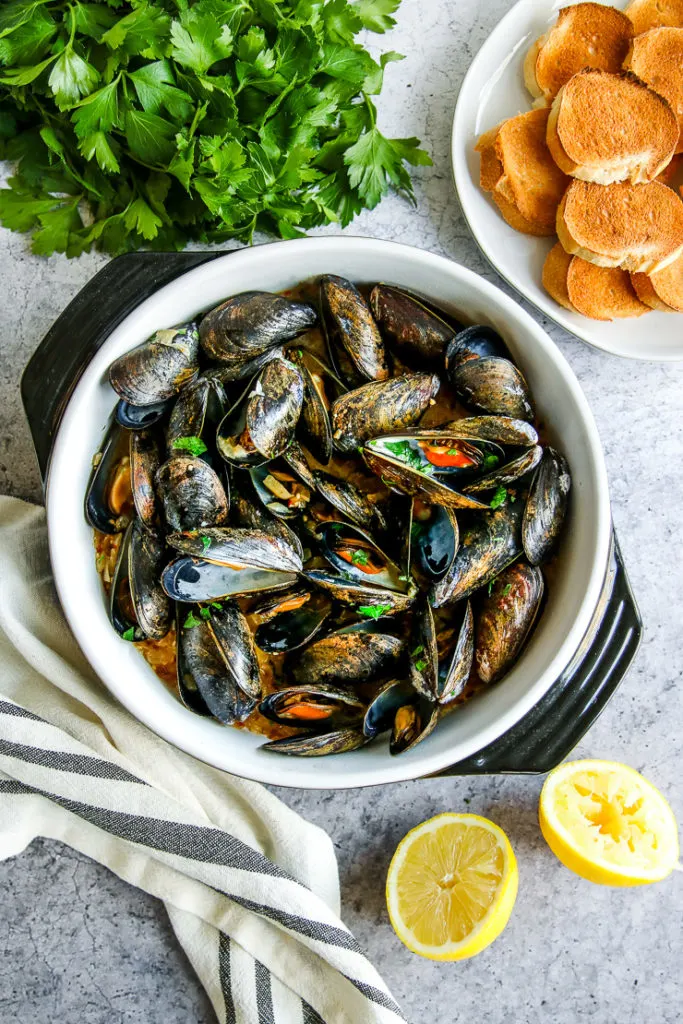 a saute pan full of garlic butter mussels next to parsley, toasted bread slices, and lemon halves