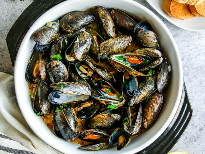 a saute pan full of garlic butter mussels next to parsley, toasted bread slices, and lemon halves