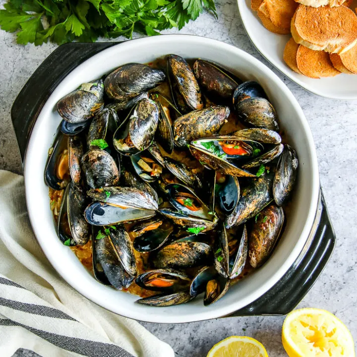 a saute pan full of garlic butter mussels next to parsley, toasted bread slices, and lemon halves