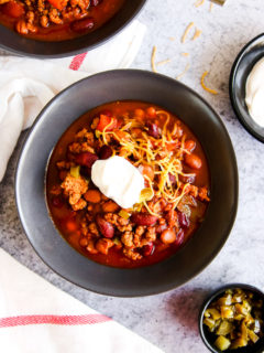 a bowl of instant pot ground turkey chili with sour cream and shredded cheese on top next to bowls of sour cream and diced jalapenos