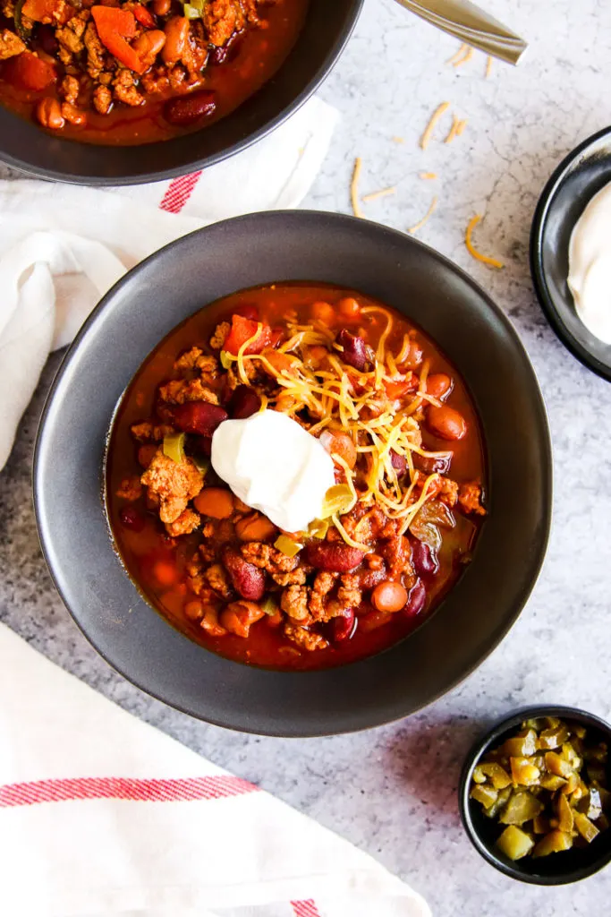 a bowl of instant pot ground turkey chili with sour cream and shredded cheese on top next to bowls of sour cream and diced jalapenos