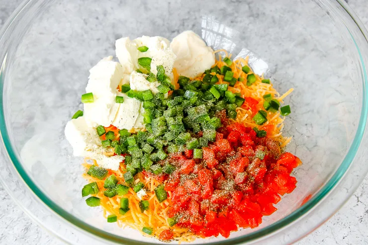 a glass bowl of shredded cheese, diced pimentos, diced jalapenos, and cream cheese with seasonings on top