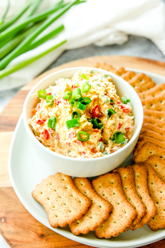 a bowl of jalapeno pimento cheese with sliced green onions on top next to crackers and full green onions