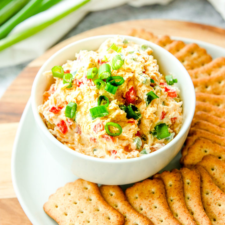 a bowl of jalapeno pimento cheese with sliced green onions on top next to crackers and full green onions