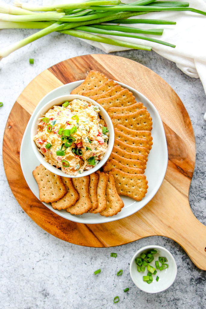 an overhead shot of a circle cutting board with a plate of crackers and bowl of jalapeno pimento cheese