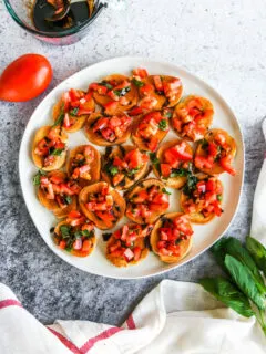 a plate of bruschetta with balsamic glaze next to a tomato, basil, and glass cup of balsamic glaze
