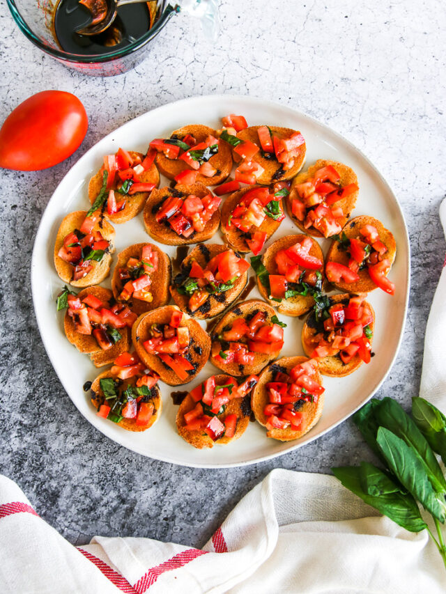 a plate of bruschetta with balsamic glaze next to a tomato, basil, and glass cup of balsamic glaze