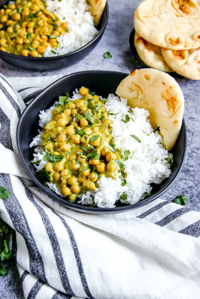 a closeup of a black bowl of chickpea coconut curry on rice with a piece of naan