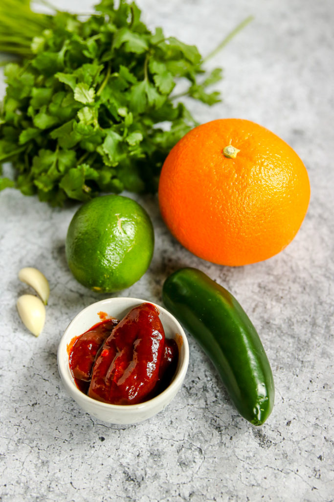 cilantro, an orange, a lime, garlic cloves, jalapeno, and chipotles in adobo