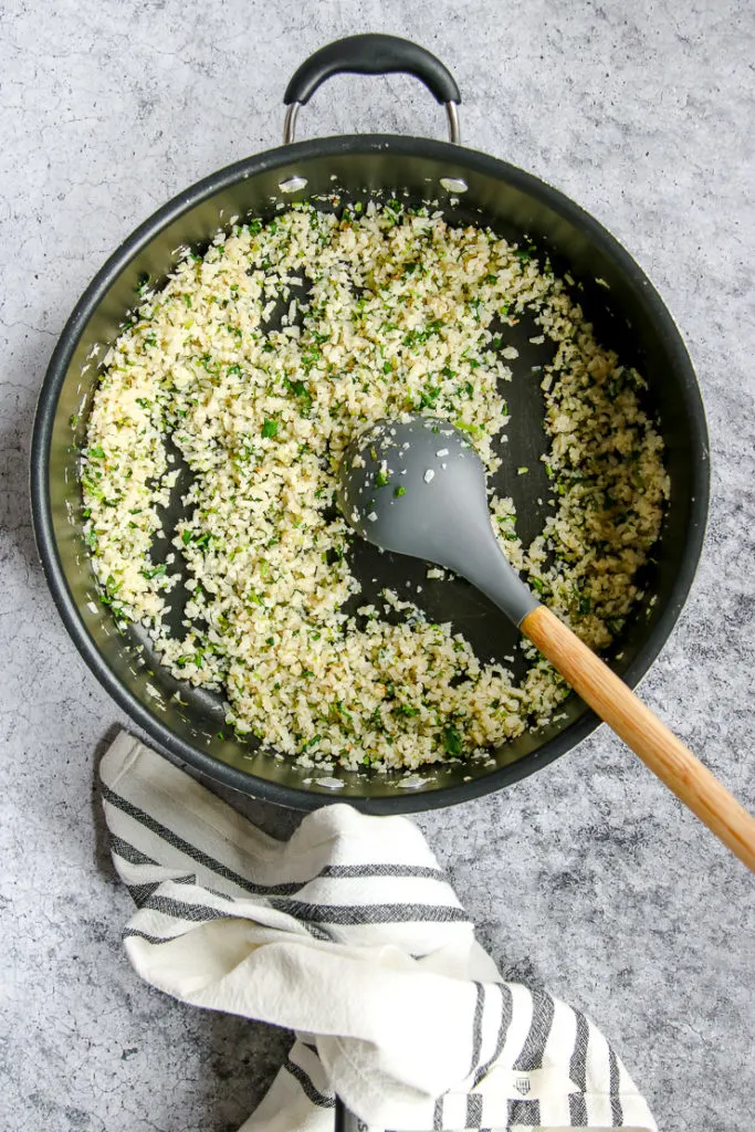 a skillet with a spoon mixing cilantro and lime into the cauliflower rice