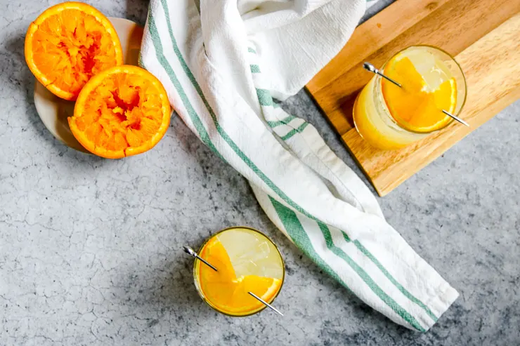 an overhead shot of squeezed oranges, two orange crushes with orange slices on cocktail picks, and a white and green striped napkin