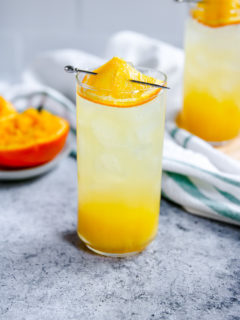 an orange crush in a highball glass with squeezed oranges behind it