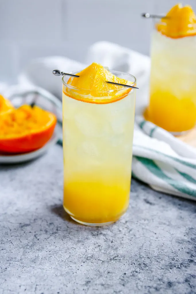 an orange crush in a highball glass with squeezed oranges behind it