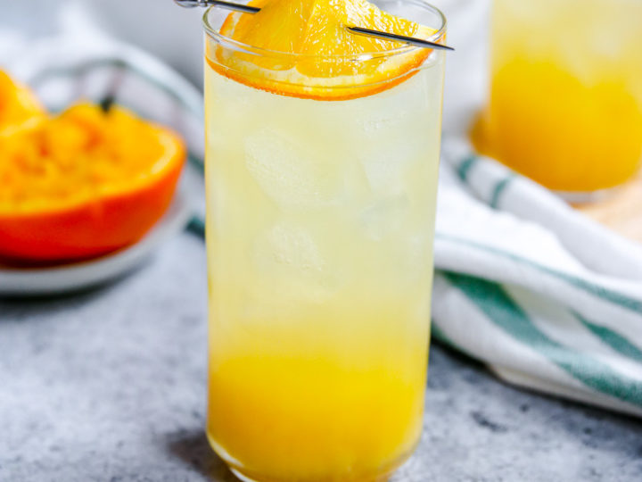 an orange crush in a highball glass with squeezed oranges behind it