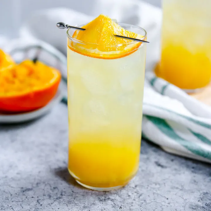 an orange crush in a highball glass with squeezed oranges behind it