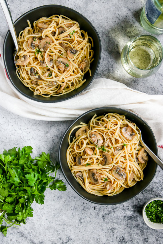 two bowls of white wine mushroom pasta next to a glass of white wine and parsley