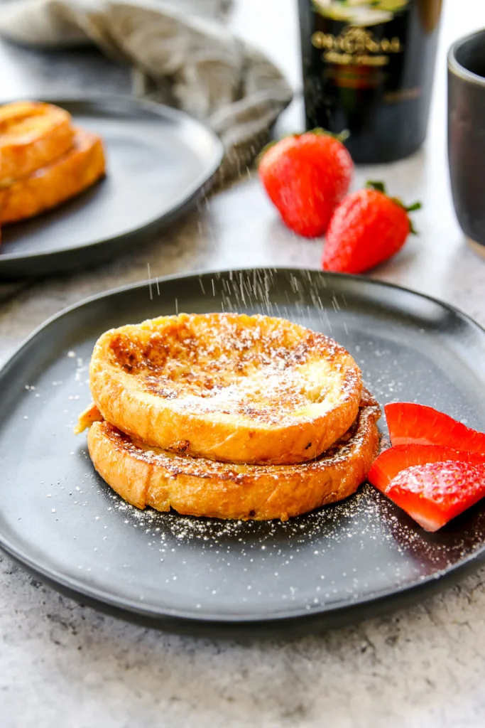 powdered sugar being sprinkled on two slices of french toast on a black plate with strawberries