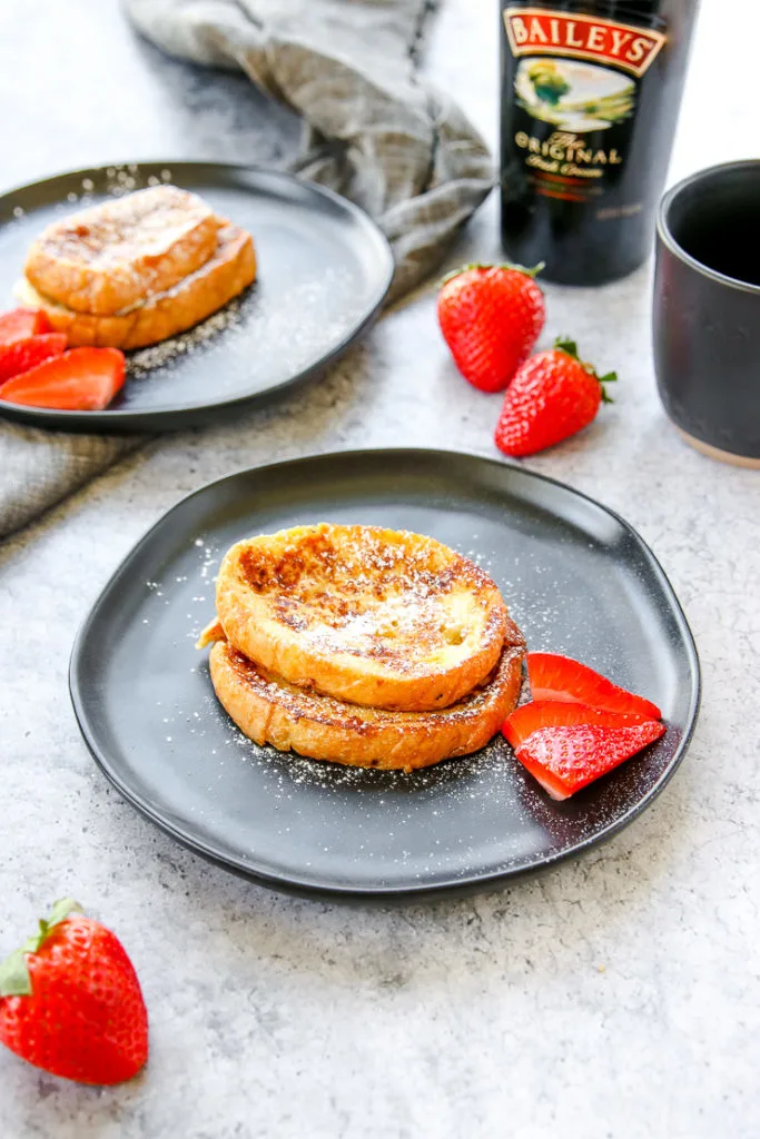 two plates of baileys french toast with strawberries, a mug of coffee, and bottle of baileys