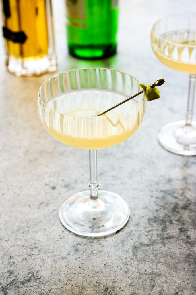 a coupe glass with elderflower gimlet in it with a gin and elderflower liquor bottle behind it