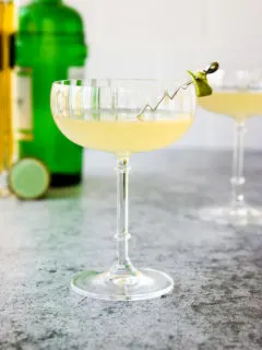 a glass of elderflower gimlet with a lime rind on a cocktail pick and green gin bottle behind it