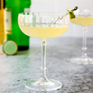 a glass of elderflower gimlet with a lime rind on a cocktail pick and green gin bottle behind it