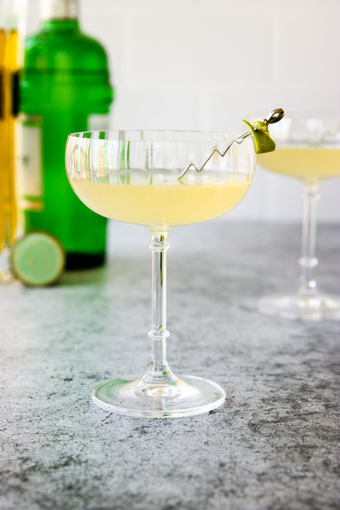 a glass of elderflower gimlet with a lime rind on a cocktail pick and green gin bottle behind it