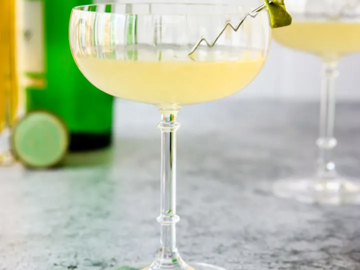 a glass of elderflower gimlet with a lime rind on a cocktail pick and green gin bottle behind it