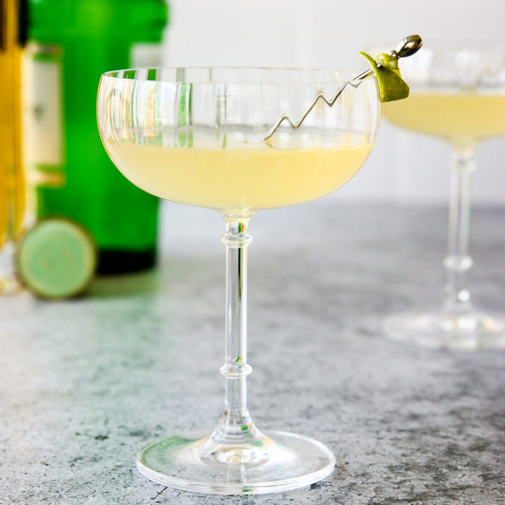 a glass of elderflower gimlet with a lime rind on a cocktail pick and green gin bottle behind it