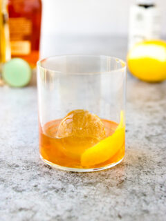 a rocks glass with a lemon peel, round ice cube, and elderflower old fashioned