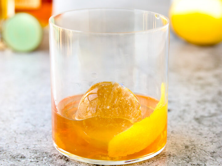 a rocks glass with a lemon peel, round ice cube, and elderflower old fashioned