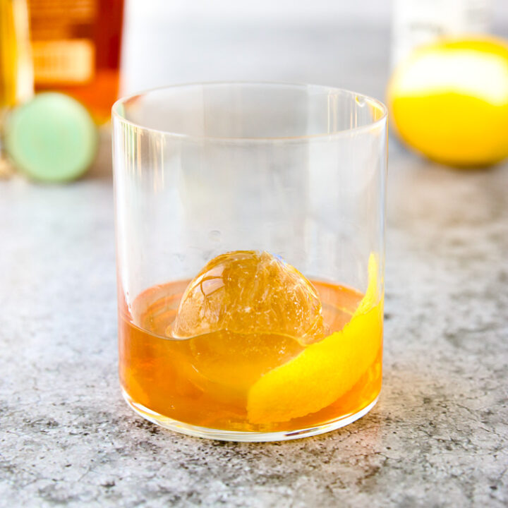 a rocks glass with a lemon peel, round ice cube, and elderflower old fashioned