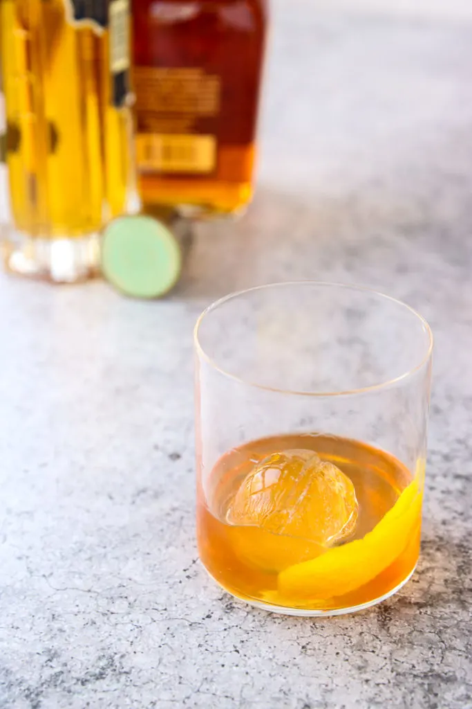 an elderflower old fashioned in a rocks glass in front of a bourbon and elderflower liquor bottle