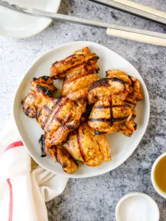 a white plate of grilled boneless skinless chicken thighs next to a napkin and grill tongs