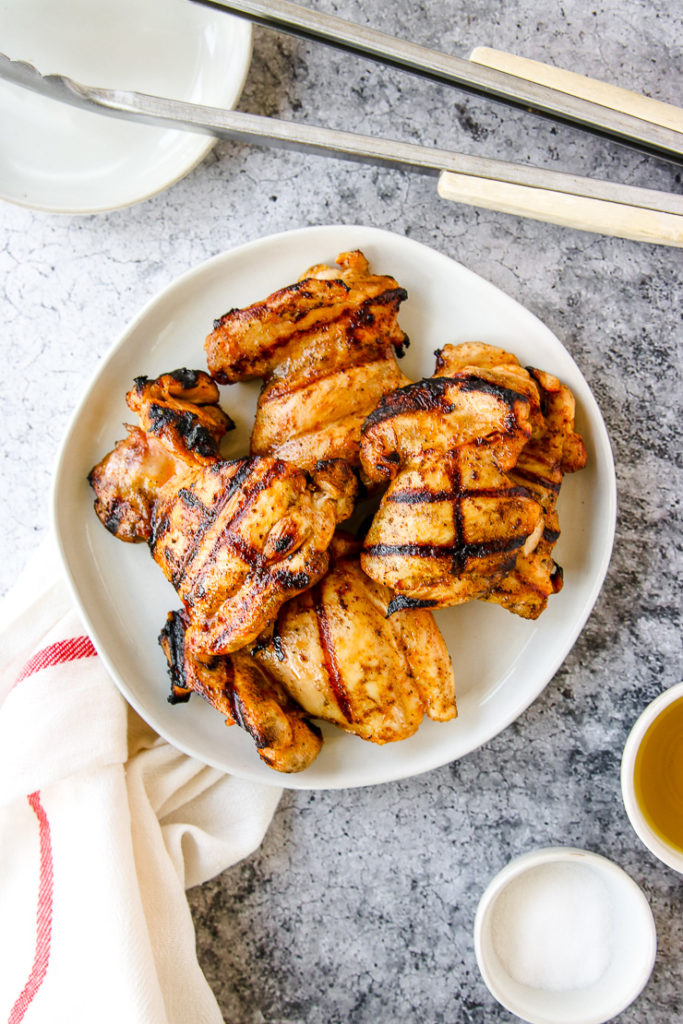 a white plate of grilled boneless skinless chicken thighs next to a napkin and grill tongs