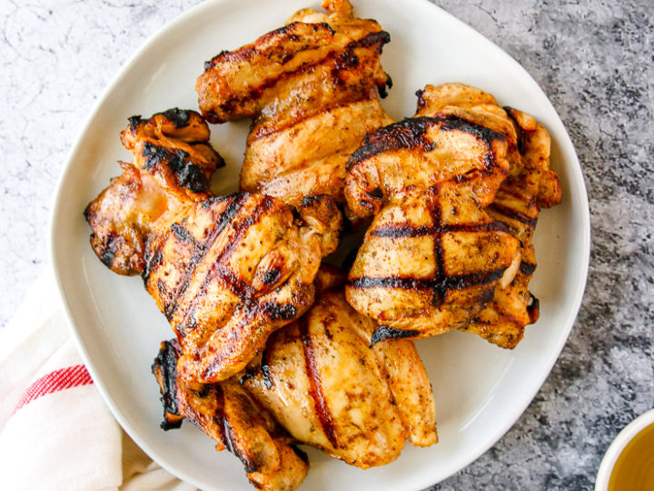 a white plate of grilled boneless skinless chicken thighs next to a napkin and grill tongs