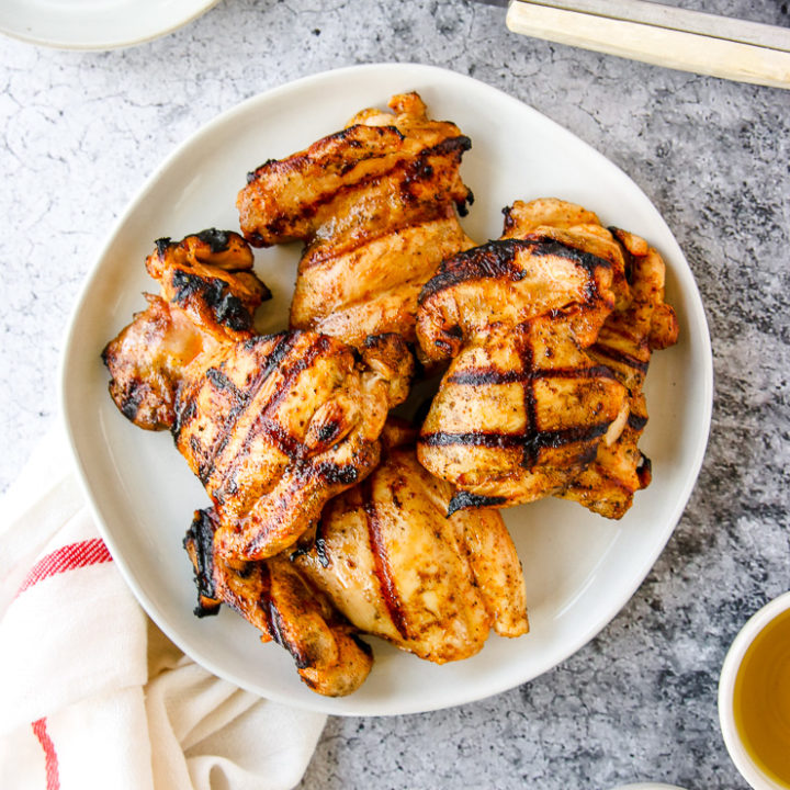 a white plate of grilled boneless skinless chicken thighs next to a napkin and grill tongs