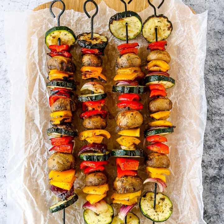 four grilled veggie skewers on wax paper on a wooden platter