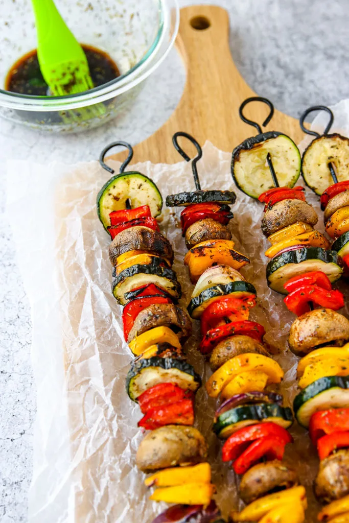 a bowl of marinade with a silicone brush next to four skewers of grilled veggies