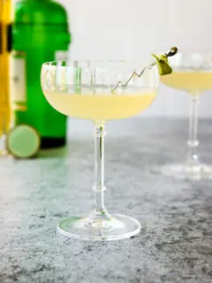 a glass of elderflower gimlet with a lime rind on a cocktail pick and green gin bottle behind it